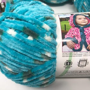 May include: A skein of bulky weight yarn in a variegated blend of bright turquoise, white, and dark grey. The yarn label features a photograph of a child wearing a hooded jacket and includes technical specifications for 5.5 mm and 6.5 mm needles.