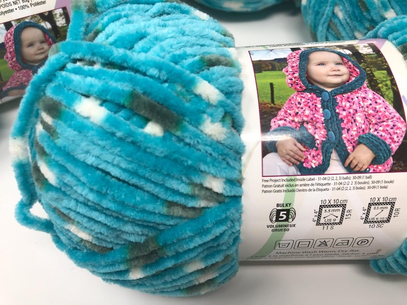 May include: A skein of bulky weight yarn in a variegated blend of bright turquoise, white, and dark grey. The yarn label features a photograph of a child wearing a hooded jacket and includes technical specifications for 5.5 mm and 6.5 mm needles.