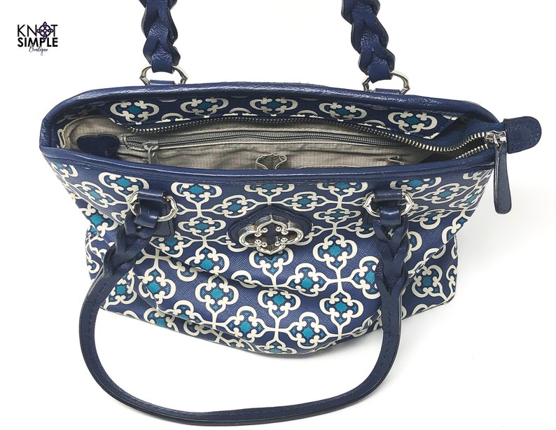 May include: A navy blue tote bag with a white and teal geometric pattern. The bag features braided handles and a silver clasp. The interior is visible, showing a zippered pocket and a gray lining. The bag has the text "Knot Simple Boutique".