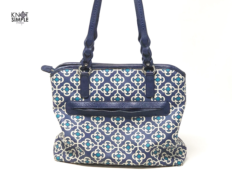 May include: A navy blue tote bag with a geometric teal and white pattern. The bag has navy blue handles and trim, a zippered top, and a front pocket. The bag has the text "KNOT SIMPLE" on the upper left corner.