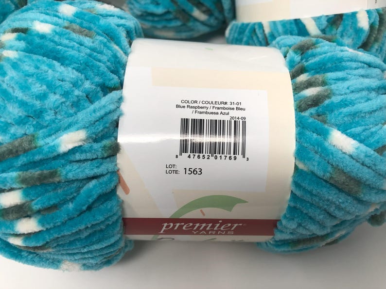 May include: A skein of plush, velvet-textured yarn in a variegated pattern of bright turquoise, white, and dark grey. A white paper label wraps around the center, displaying the text Premier Yarns, color 31-01 Blue Raspberry, and a barcode.