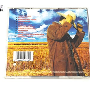 May include: A CD case featuring a man in a brown coat and hat, set against a backdrop of a wheat field and blue sky. The CD case has a tracklist with titles like "Big Big Love" and "Trail of Broken Hearts".