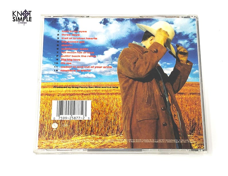 May include: A CD case featuring a man in a brown coat and hat, set against a backdrop of a wheat field and blue sky. The CD case has a tracklist with titles like "Big Big Love" and "Trail of Broken Hearts".