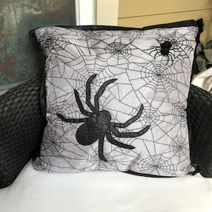 May include: A square throw pillow featuring a light gray fabric base with a black spiderweb pattern printed across the surface. A large, black, textured spider is centered on the front, with a smaller black spider attached near the top right corner.