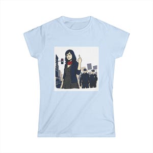 May include: A light blue t-shirt featuring a graphic illustration of a person with dark hair wearing a dark cardigan over a polka dot dress and a red scarf, gesturing with a middle finger toward two police officers in a city street setting.