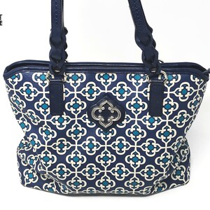 May include: A navy blue tote bag with a white and teal geometric pattern. The bag has navy blue handles and trim, with a silver-colored decorative emblem on the front. The bag is designed with a zipper closure.