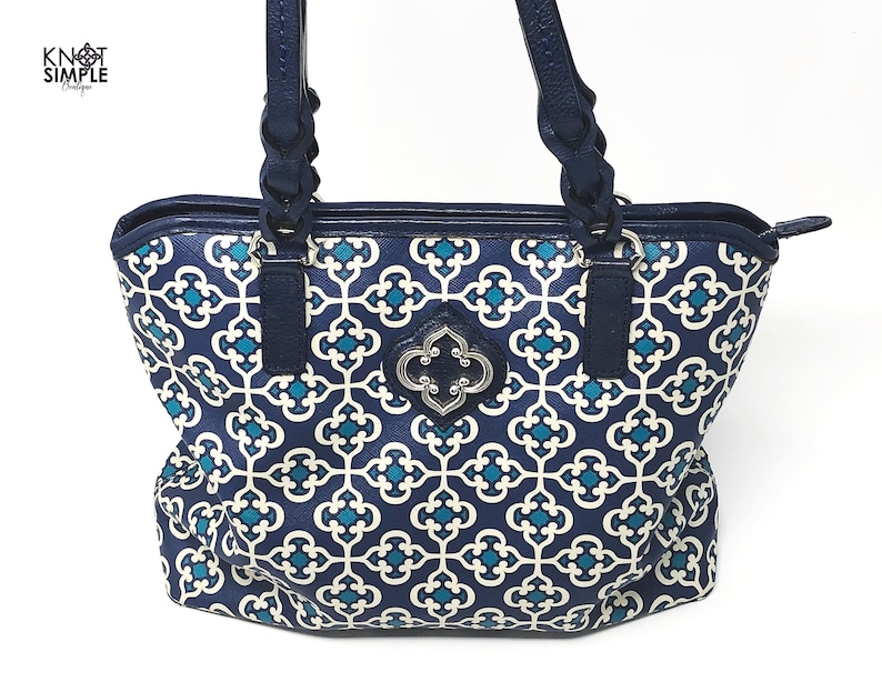 May include: A navy blue tote bag with a white and teal geometric pattern. The bag has navy blue handles and trim, with a silver-colored decorative emblem on the front. The bag is designed with a zipper closure.