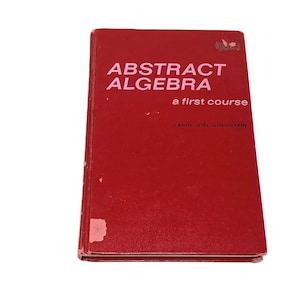 May include: A red book titled "ABSTRACT ALGEBRA" with the subtitle "a first course" and the author's name, Larry Joel Goldstein. The book has a worn cover with some damage on the bottom left corner.