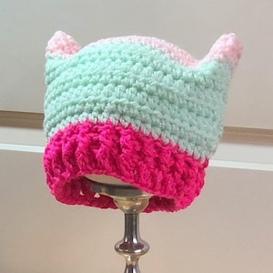 May include: A crocheted beanie with cat-ear points, featuring a bright hot pink ribbed brim and a light mint green upper section. The yarn has a subtle metallic shimmer throughout, and the hat is constructed with a thick, textured stitch pattern.