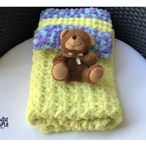 May include: A brown plush teddy bear with a tan ribbon around its neck sits on a folded yellow textured blanket. A smaller, folded blanket with a purple, blue, and yellow variegated pattern rests behind the bear. The text KNOT SIMPLE Boutique is visible.