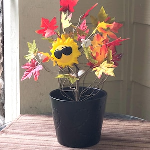 May include: A whimsical decorative plant in a black pot. The plant features a yellow sun wearing sunglasses, surrounded by colorful artificial leaves in shades of red, orange, and yellow. The pot sits on a brown and tan striped surface.