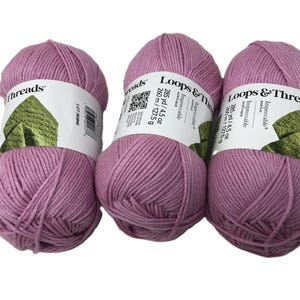 May include: Three balls of Loops & Threads Impeccable acrylic yarn in a dusty rose color. Each ball has a label with product information, including 285 yards / 4.5 oz. The yarn is suitable for various knitting and crochet projects.