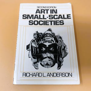 May include: A book titled "Art in Small-Scale Societies" by Richard L. Anderson. The cover features a black and white image of a tribal mask. The book is a second edition and has a white cover with black text.