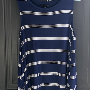 May include: Navy blue sleeveless top with horizontal white stripes. The top is hanging on a blue hanger. The brand name "SIGNATURE STUDIO" is printed on the top.