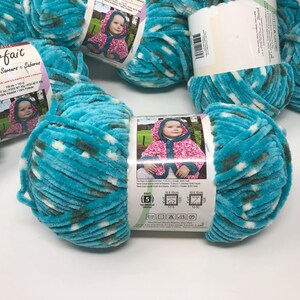 May include: A skein of turquoise, white, and dark gray variegated chenille yarn labeled Parfait. The paper label features a photograph of a child in a hooded jacket, product specifications, and care instructions for 100% polyester material.