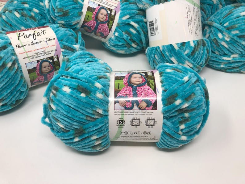 May include: A skein of turquoise, white, and dark gray variegated chenille yarn labeled Parfait. The paper label features a photograph of a child in a hooded jacket, product specifications, and care instructions for 100% polyester material.