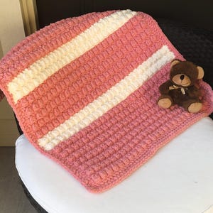 May include: A chunky crochet blanket featuring a textured bobble stitch pattern in alternating stripes of coral pink and cream white. A small, seated brown plush teddy bear with a cream-colored ribbon around its neck rests on the blanket surface.