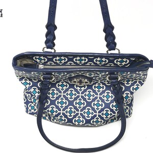 May include: A navy blue tote bag with a white and teal geometric pattern. The bag has braided navy blue handles and a silver clasp. The interior lining features a similar pattern. The bag is open, revealing the interior.