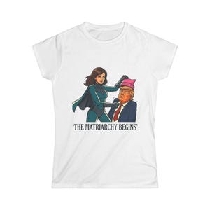 May include: A white short-sleeved t-shirt featuring a graphic illustration of a superhero figure placing a pink knit hat on a man in a suit and red tie. Below the illustration, the text reads THE MATRIARCHY BEGINS in black capital letters.