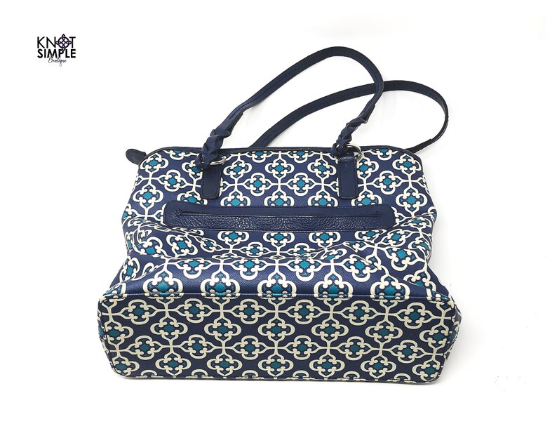 May include: Navy blue tote bag with a geometric pattern in teal and white. The bag features a zippered pocket and braided handles. The brand name "Knot Simple" is visible in the upper left corner.