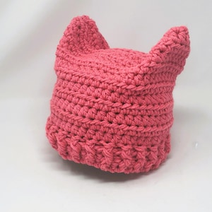 May include: A pink crocheted hat with cat ears. The hat is made with a single crochet stitch and has a simple design.