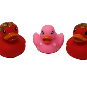 May include: Three rubber duck toys. Two are red with orange beaks and decorative headbands. The center duck is pink with white heart accents and an orange beak. All ducks have black eyes and are on a white background.