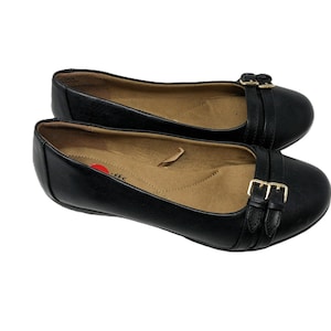 May include: A pair of black ballet flats featuring a rounded toe and two decorative straps across the front, each secured with a small gold-toned metal buckle. The interior of the shoes is tan with a red circular logo on the heel insole.