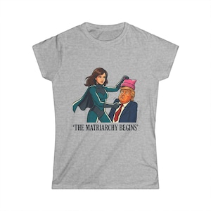 May include: A heather gray t-shirt featuring a graphic illustration of a superhero figure placing a pink knit hat on a man in a suit and red tie. Below the illustration, the text reads, THE MATRIARCHY BEGINS, in a bold, dark serif font.