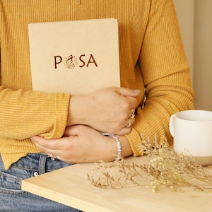 May include: A light brown rectangular notebook with the text POSA printed in dark brown, featuring a butterfly graphic in place of the letter O. The notebook is held against a mustard yellow ribbed long-sleeve shirt and blue denim jeans.