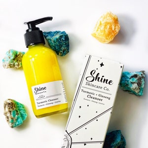 Organic skincare turmeric cleanser in clear glass bottle with pump dispenser. Face wash is bright yellow color and photographed on white background with white shine skincare box. Skincare products surrounded by blue and yellow crystals.