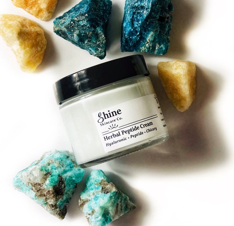 Organic skincare anti-aging face moisturizer in clear glass jar with black lid. Face cream is a very pale blue color from blue tansy oil. Photo of jar on white background surrounded by blue and yellow crystal gemstones.