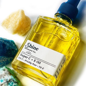 May include: A square glass bottle of Shine Skincare Co. Super C + E Oil with a blue dropper top. The bottle contains a yellow liquid and has a white label with product information. The bottle is surrounded by colorful rocks.