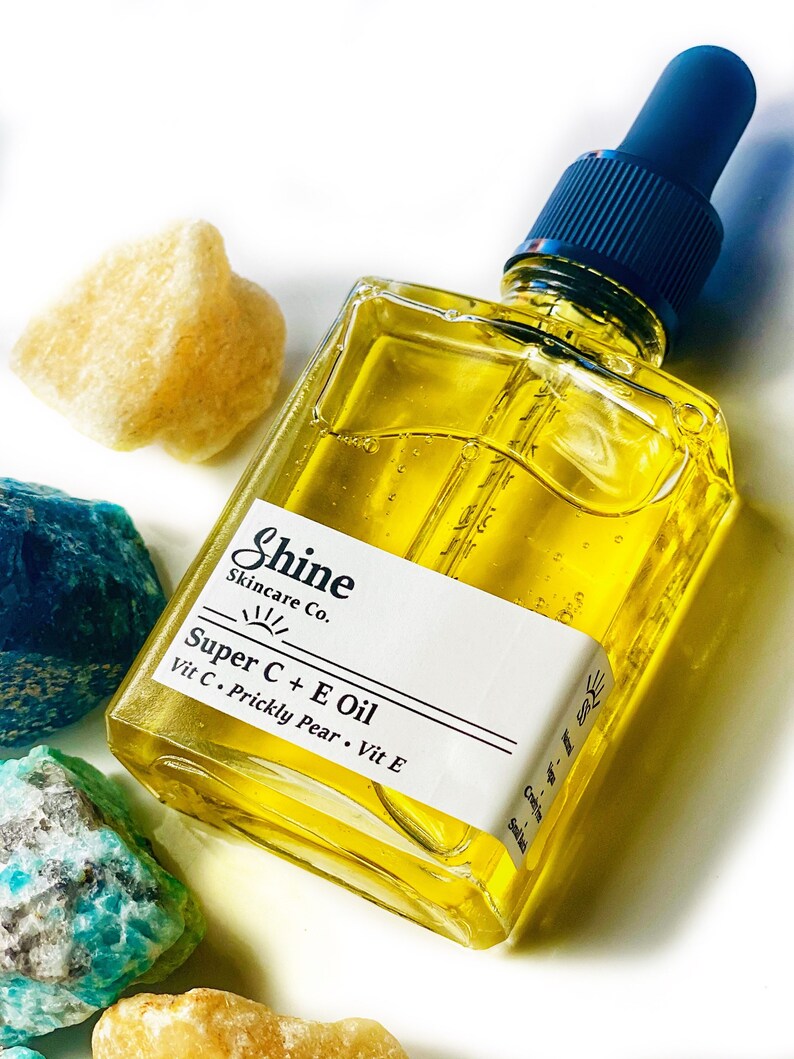 May include: A square glass bottle of Shine Skincare Co. Super C + E Oil with a blue dropper top. The bottle contains a yellow liquid and has a white label with product information. The bottle is surrounded by colorful rocks.