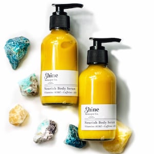 Puede incluir: Dos botellas de vidrio de Shine Skincare Co. Nourish Body Serum con tapas de bomba negras, llenas de un l&iacute;quido amarillo. Las botellas est&aacute;n rodeadas de piedras preciosas coloridas y en bruto sobre un fondo blanco. La etiqueta incluye Vitaminas AC&E y Cafe&iacute;na.