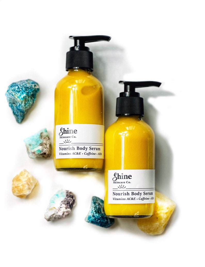 Puede incluir: Dos botellas de vidrio de Shine Skincare Co. Nourish Body Serum con tapas de bomba negras, llenas de un l&iacute;quido amarillo. Las botellas est&aacute;n rodeadas de piedras preciosas coloridas y en bruto sobre un fondo blanco. La etiqueta incluye Vitaminas AC&E y Cafe&iacute;na.