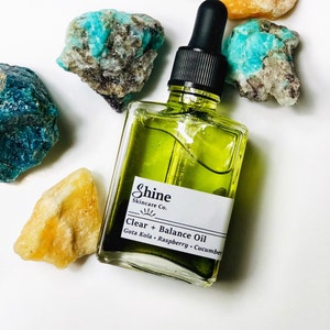 May include: A clear glass bottle with a dropper filled with green liquid. The bottle has a white label with the text "Shine Skincare Co. Clear + Balance Oil Gota Kola - Raspberry - Cucumber".