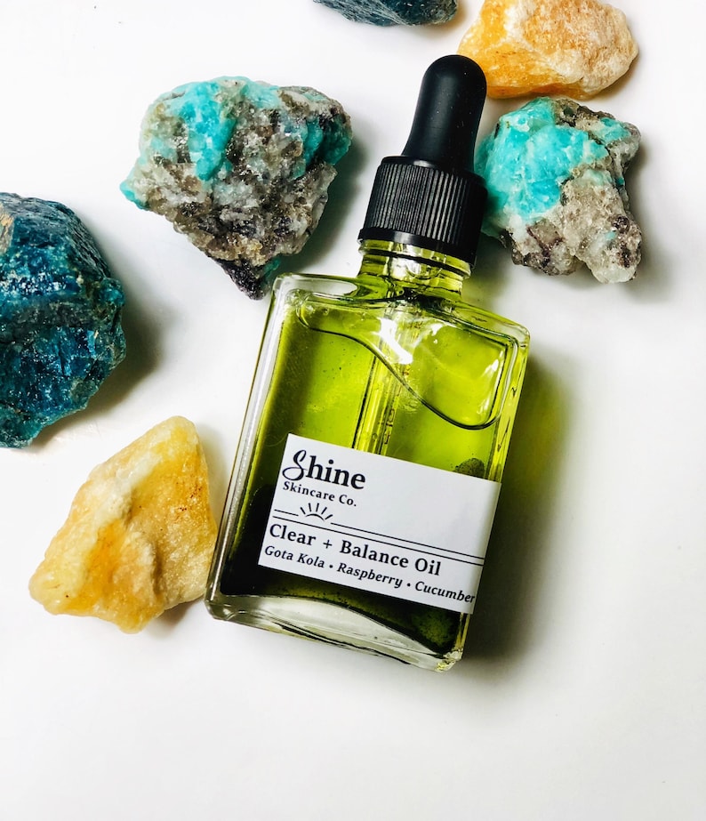 May include: A clear glass bottle with a dropper filled with green liquid. The bottle has a white label with the text "Shine Skincare Co. Clear + Balance Oil Gota Kola - Raspberry - Cucumber".