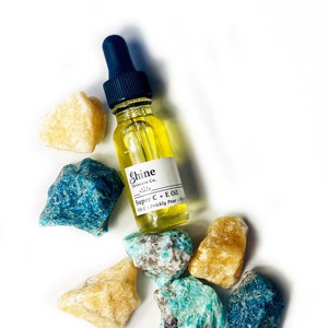 May include: A clear glass bottle of Shine Skincare Co. Super C + E Oil with a blue dropper, surrounded by yellow and blue-green gemstones. The bottle has a white label with product information.