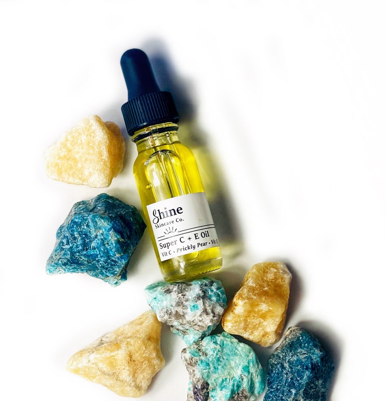 May include: A clear glass bottle of Shine Skincare Co. Super C + E Oil with a blue dropper, surrounded by yellow and blue-green gemstones. The bottle has a white label with product information.