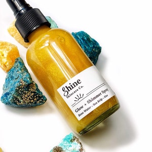 May include: A clear glass spray bottle filled with a golden liquid, labeled "Shine Skincare Co. Glow + Shimmer Spray." The bottle is surrounded by colorful, rough-cut gemstones in shades of blue, yellow, and turquoise. The spray bottle has a black nozzle.