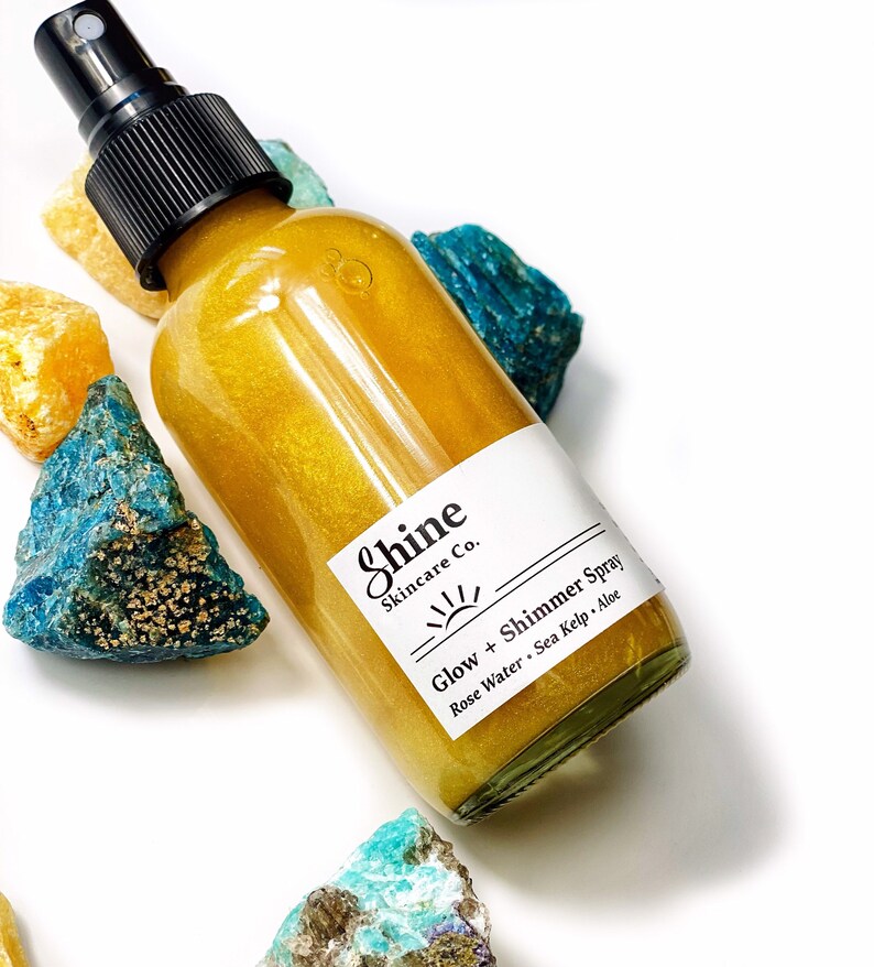 May include: A clear glass spray bottle filled with a golden liquid, labeled "Shine Skincare Co. Glow + Shimmer Spray." The bottle is surrounded by colorful, rough-cut gemstones in shades of blue, yellow, and turquoise. The spray bottle has a black nozzle.