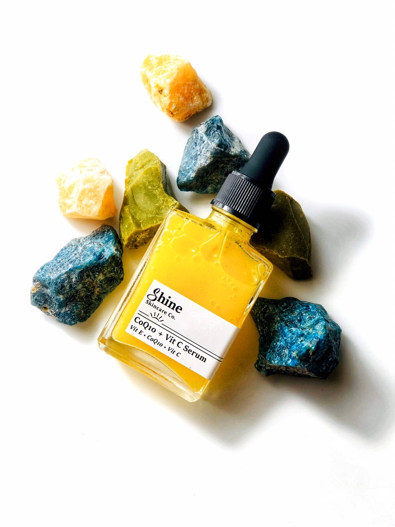 Organic skincare product vitamin c serum in clear square glass bottle. Serum is pale yellow color and thin consistency. Photographed on white background with blue, yellow and green crystals surrounding serum.