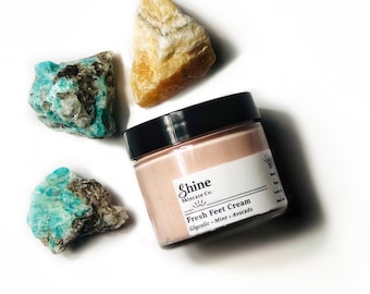 May include: A white jar with a black lid and a label that reads "Shine Skincare Co. Fresh Feet Cream Glycolic - Mint - Avocado". The jar is surrounded by three light blue and white crystals.