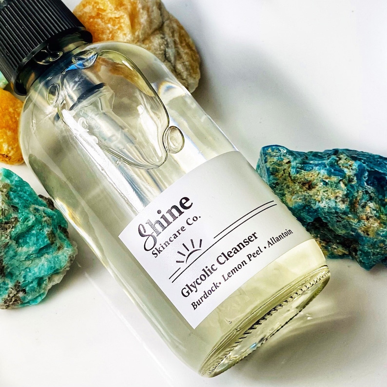 Pu&ograve; includere: Una bottiglia di vetro trasparente con un tappo a pompa nero e un'etichetta bianca che recita "Shine Skincare Co. Glycolic Cleanser Burdock, Lemon Peel, Allantoin". La bottiglia &egrave; circondata da pietre colorate.