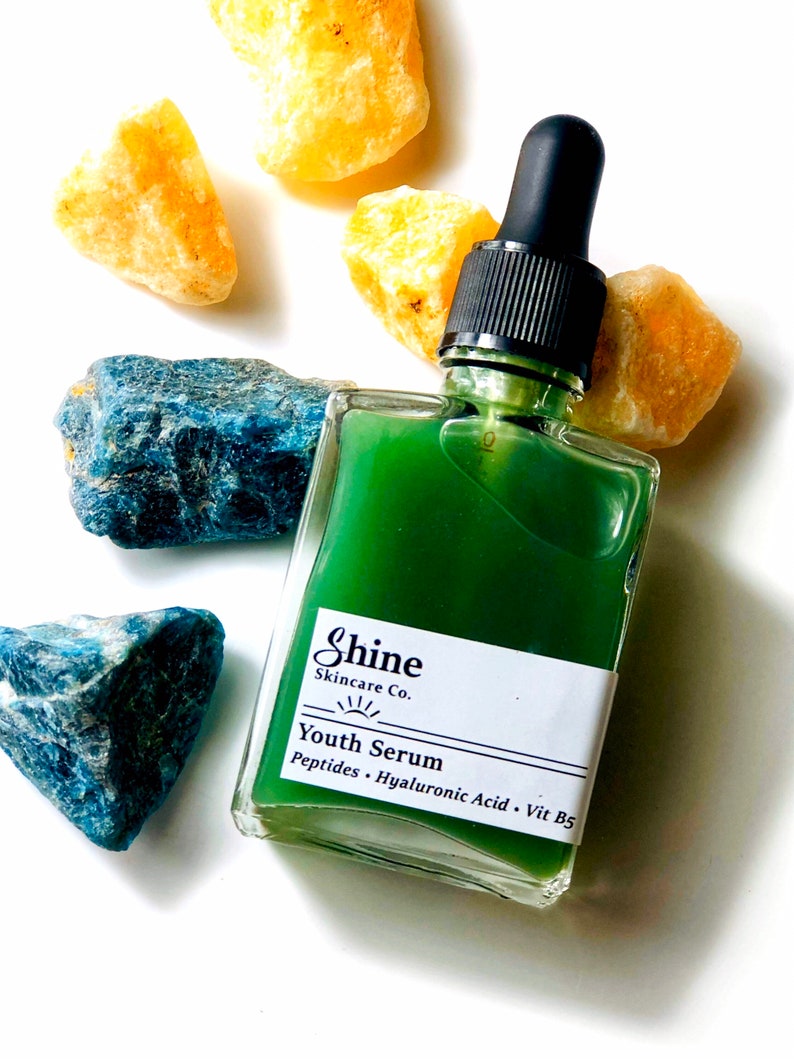 Organic skincare, natural facial serum in clear square glass with black dropper top. Anti-aging face serum is green color photographed on white background with yellow and blue crystal gemstones surrounded facial care bottle.