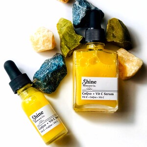 Organic skincare vitamin c serum in clear square glass bottle with smaller Boston round clear glass bottle. Serum is pale yellow color and thin consistency. Photographed on white background with blue, yellow and green crystals surrounding serum.