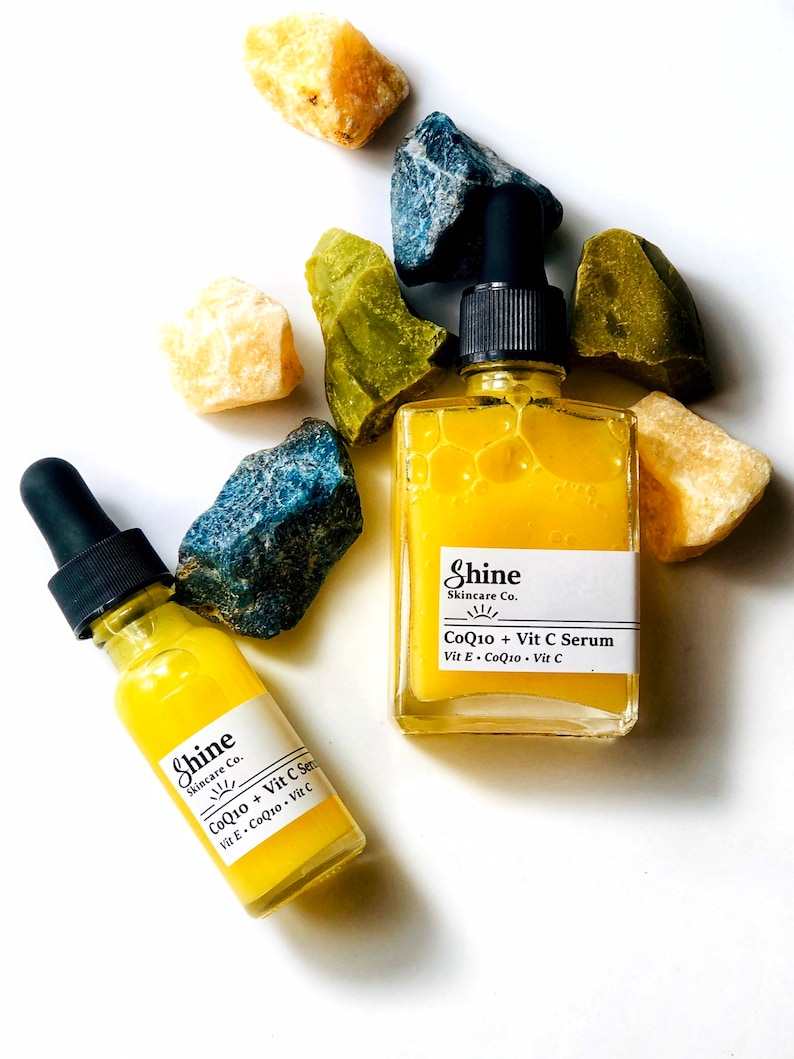 Organic skincare vitamin c serum in clear square glass bottle with smaller Boston round clear glass bottle. Serum is pale yellow color and thin consistency. Photographed on white background with blue, yellow and green crystals surrounding serum.