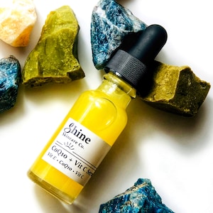 May include: A yellow glass bottle with a black dropper top and a white label that reads "Shine Skincare Co. CoQ10 + Vit C Serum Vit E. CoQ10. Vit C" surrounded by blue and green stones.