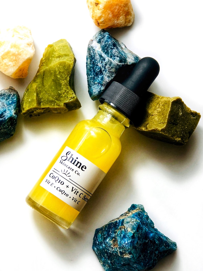 May include: A yellow glass bottle with a black dropper top and a white label that reads "Shine Skincare Co. CoQ10 + Vit C Serum Vit E. CoQ10. Vit C" surrounded by blue and green stones.