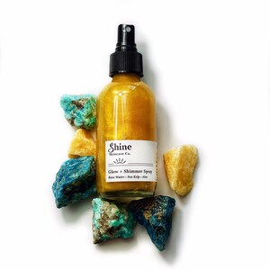 May include: A clear glass bottle filled with a golden liquid, labeled "Shine Skincare Co. Glow + Shimmer Spray." The bottle is surrounded by colorful, rough-cut gemstones in shades of blue, turquoise, and yellow. The spray bottle has a black nozzle.